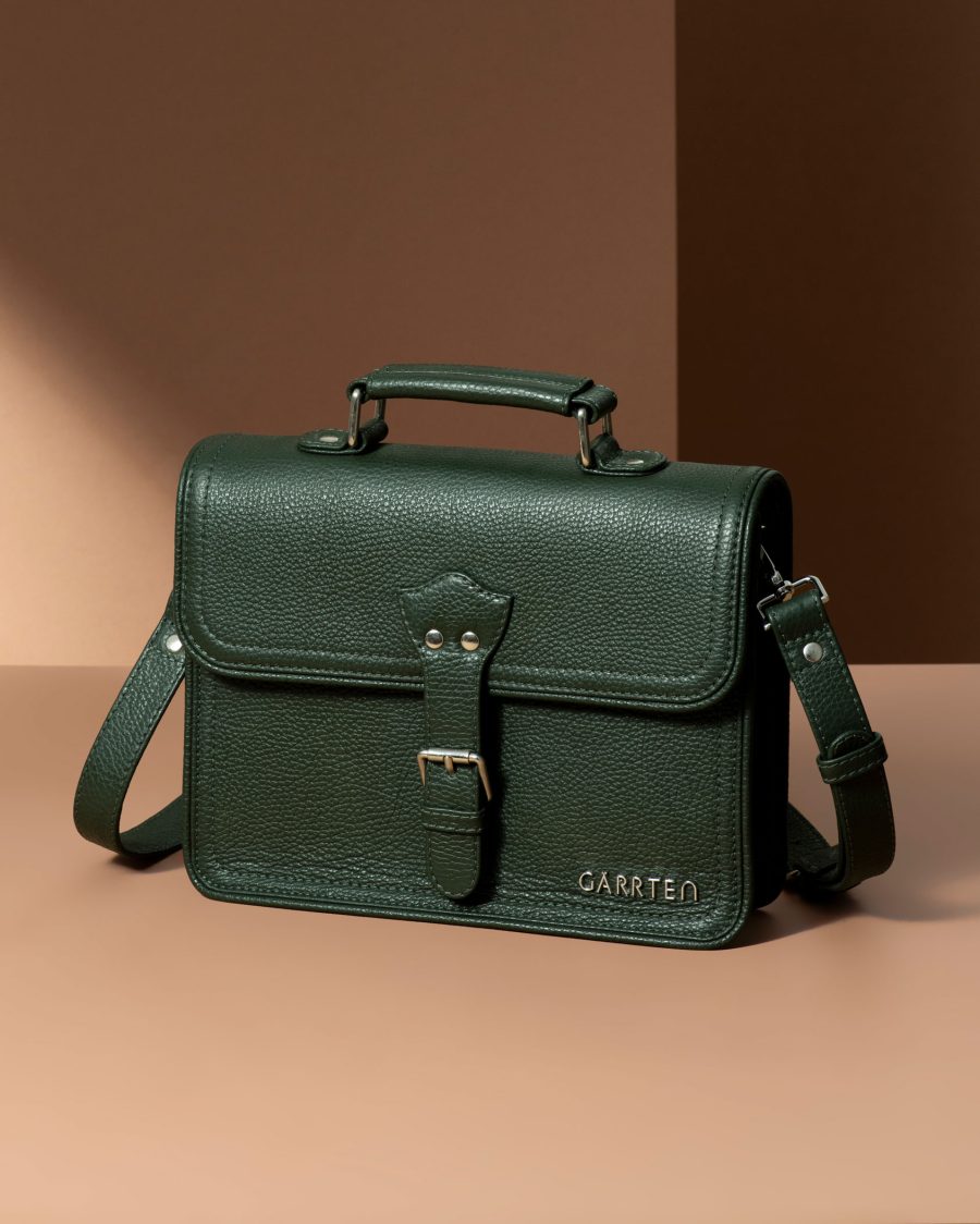Green leather satchel bag with buckle strap and shoulder sling on beige background.