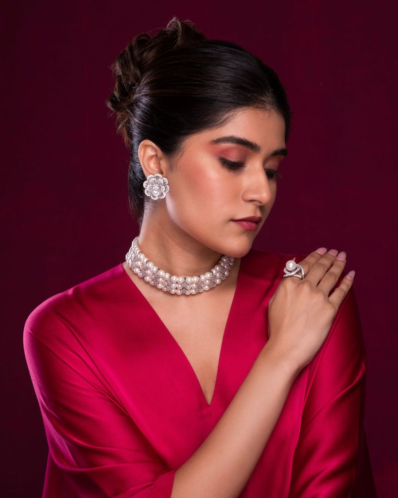 Model wearing pearl choker, floral earrings, and statement ring on burgundy background.